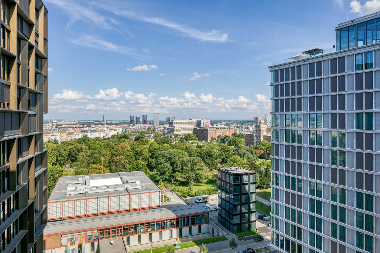 A high-rise building with a modern glass facade, surrounded by greenery and other buildings in the background.