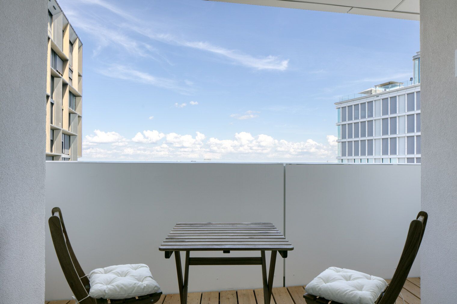 Ein Balkon mit einem Holztisch und zwei gepolsterten Stühlen, mit Blick auf eine Stadtlandschaft mit Gebäuden und blauem Himmel.