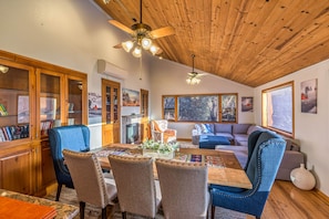 A spacious living area with a wooden ceiling, a dining table with chairs, a sofa, and a large window providing a view of the outdoors.