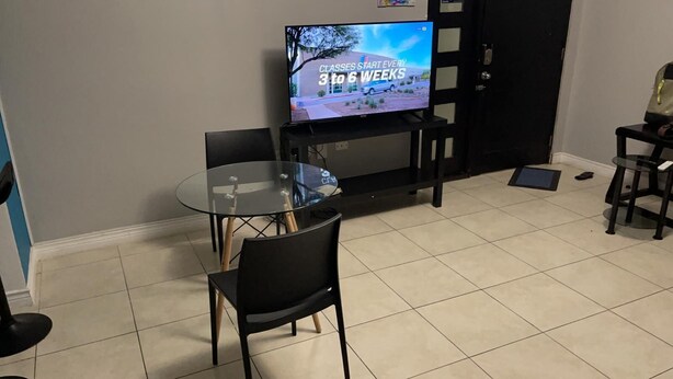 Un salon avec une table en verre, deux chaises noires, un téléviseur affichant un message concernant les cours, et un meuble noir.
