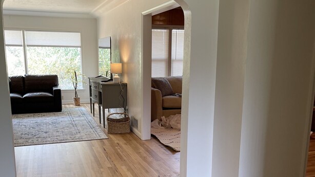 Ein geräumiger Wohnbereich mit Holzboden, einem dunkelbraunen Sofa, einem kleinen Tisch mit Lampe und einem Fenster mit Jalousien.