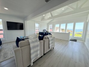 A spacious living room with a large sectional sofa, a flat-screen TV mounted on the wall, and large windows offering a view of the outdoors.