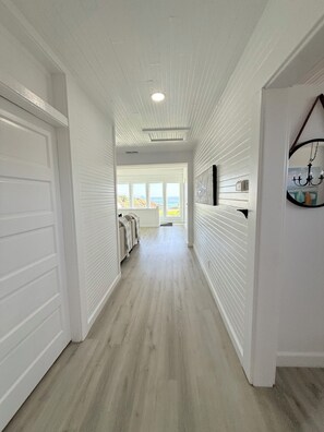 A hallway with white walls and a wooden floor, leading to a door with a view of the outdoors.