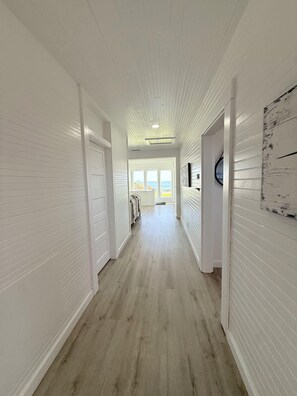A hallway with white walls and wooden floors, leading to a room with a view of the ocean.