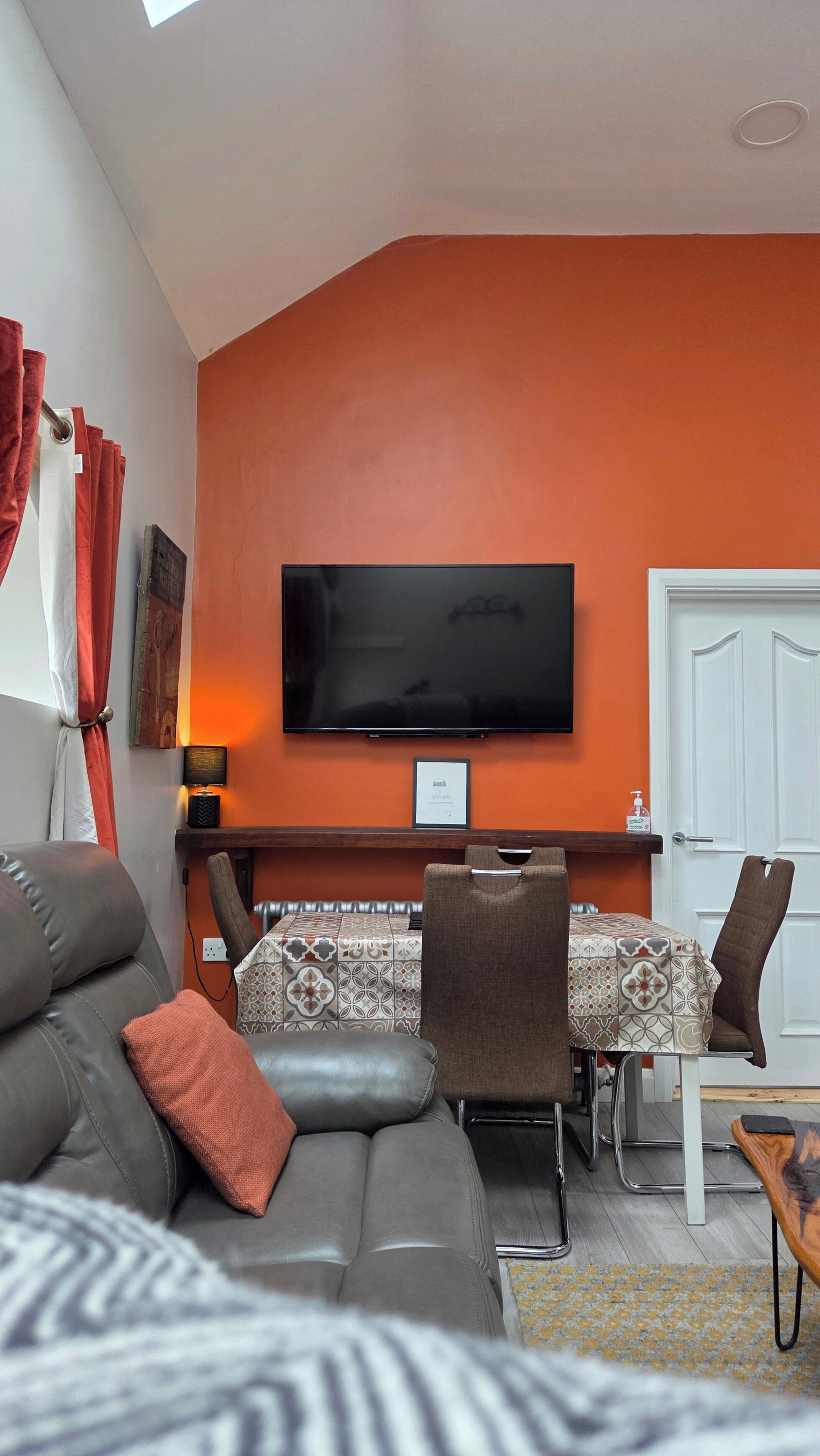 A living room with a sofa, a TV mounted on the wall, a table with chairs, and a door.