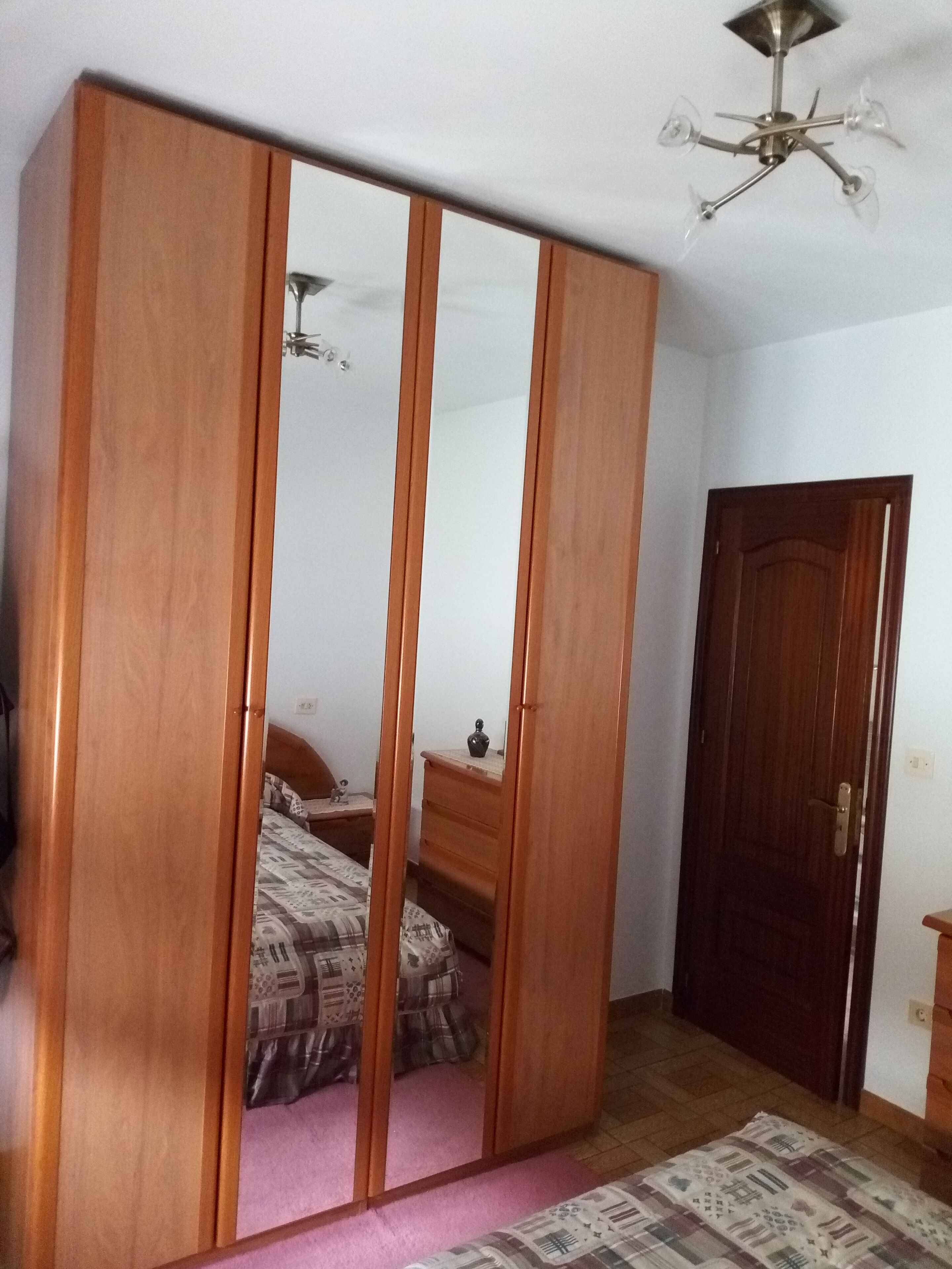 Une chambre avec une armoire en bois, un lit avec une literie à motifs, une commode en bois et une porte.