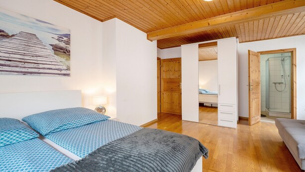 Une chambre avec un plafond en bois, un lit avec une literie bleue et grise, une porte en bois donnant sur une salle de bain et un tableau accroché au mur.