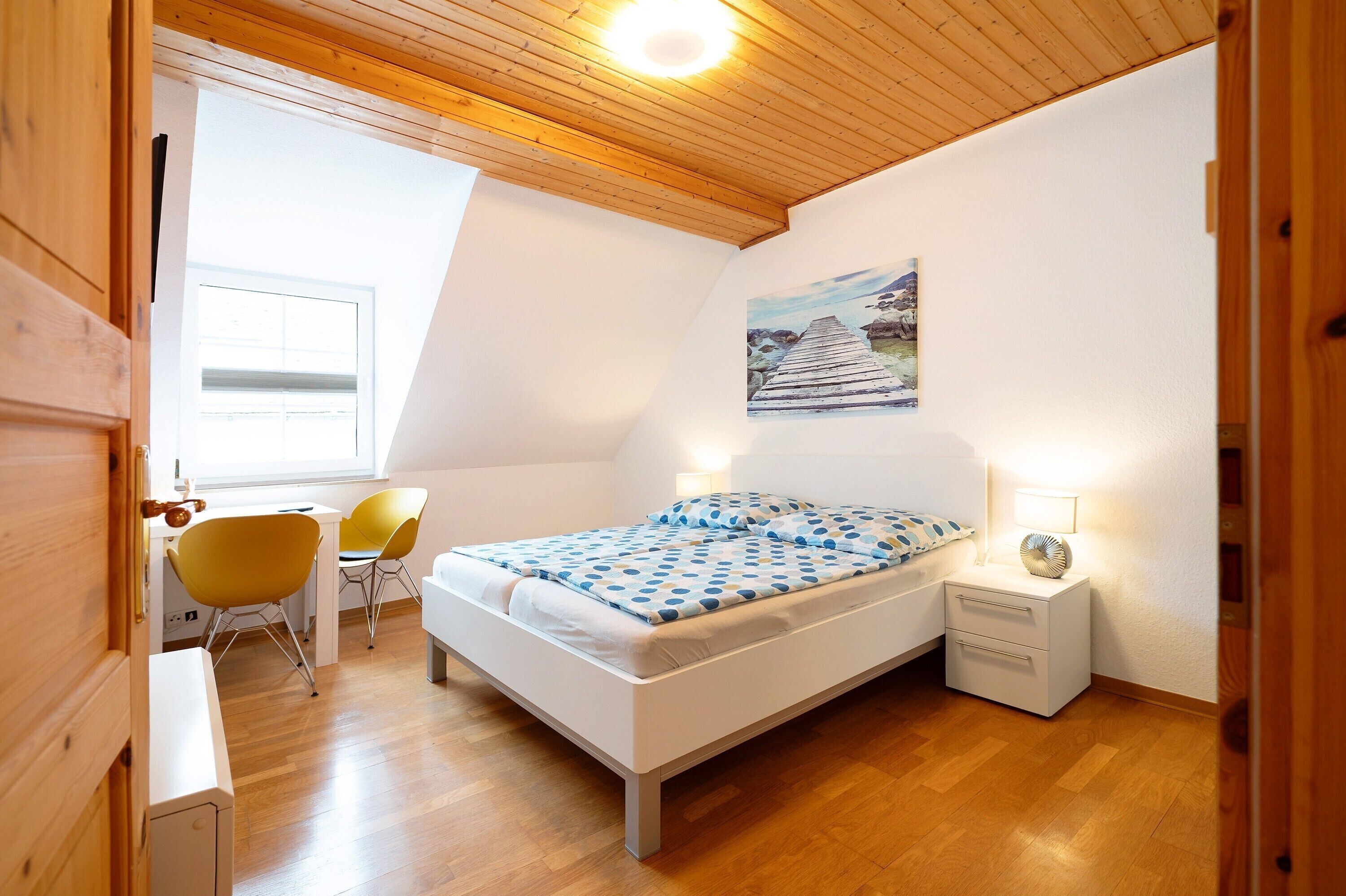 Um quarto com uma cama, uma escrivaninha, duas cadeiras, uma mesa de cabeceira e um quadro na parede.