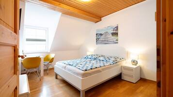 Um quarto com uma cama, uma escrivaninha, duas cadeiras, uma mesa de cabeceira e um quadro na parede.