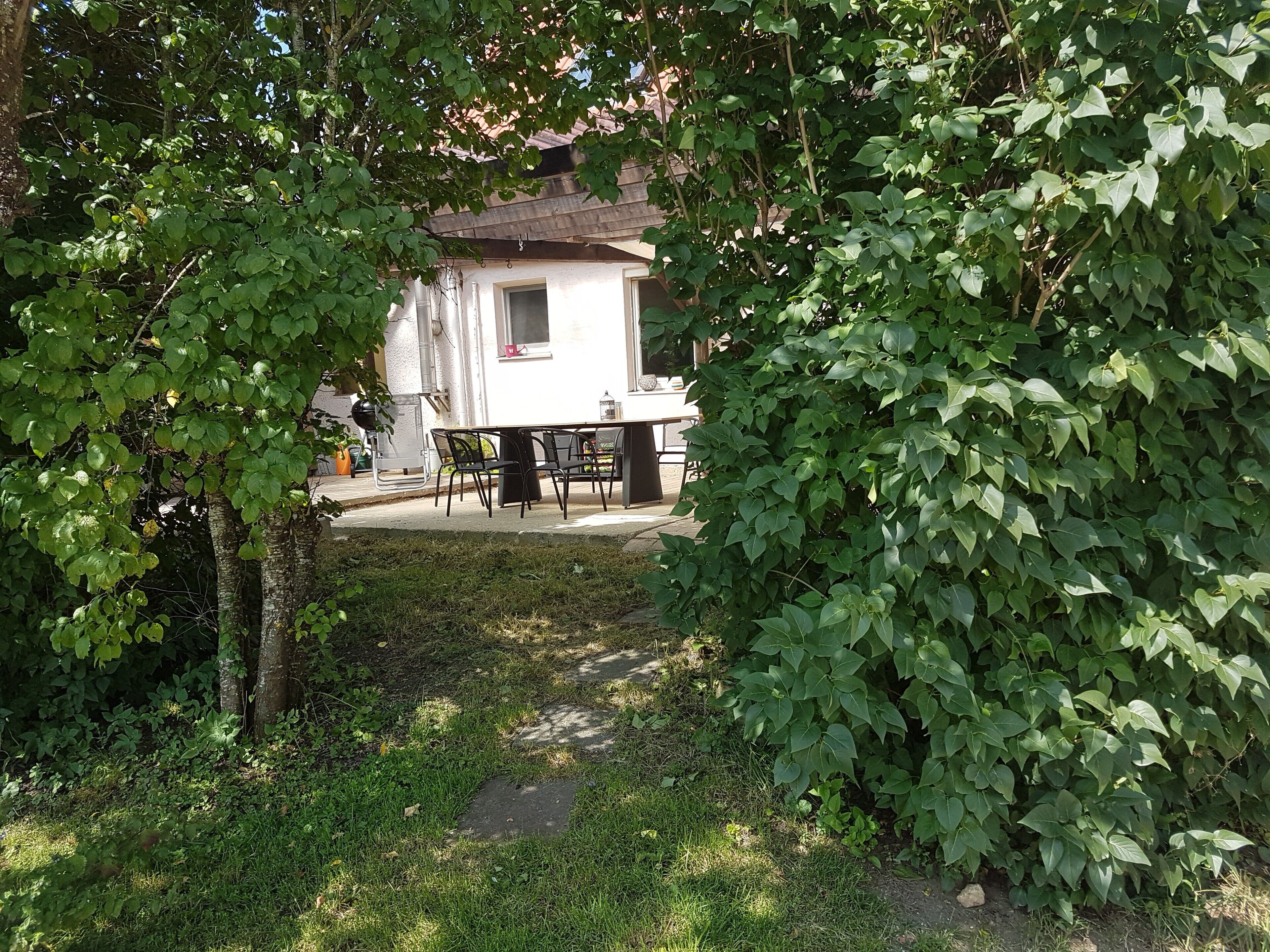 Un bâtiment blanc avec un toit en tuiles, une table entourée de chaises et un jardin avec de la pelouse et des arbres.
