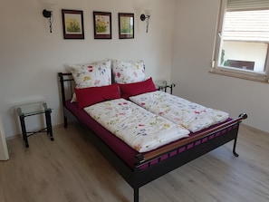 A bed with floral bedding, a red pillow, and a wooden headboard. There are two bedside tables, a window with blinds, and wall-mounted lamps.