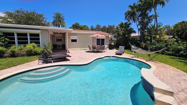 Una piscina con borde de baldosas, un patio cubierto con mobiliario exterior y una hamaca.