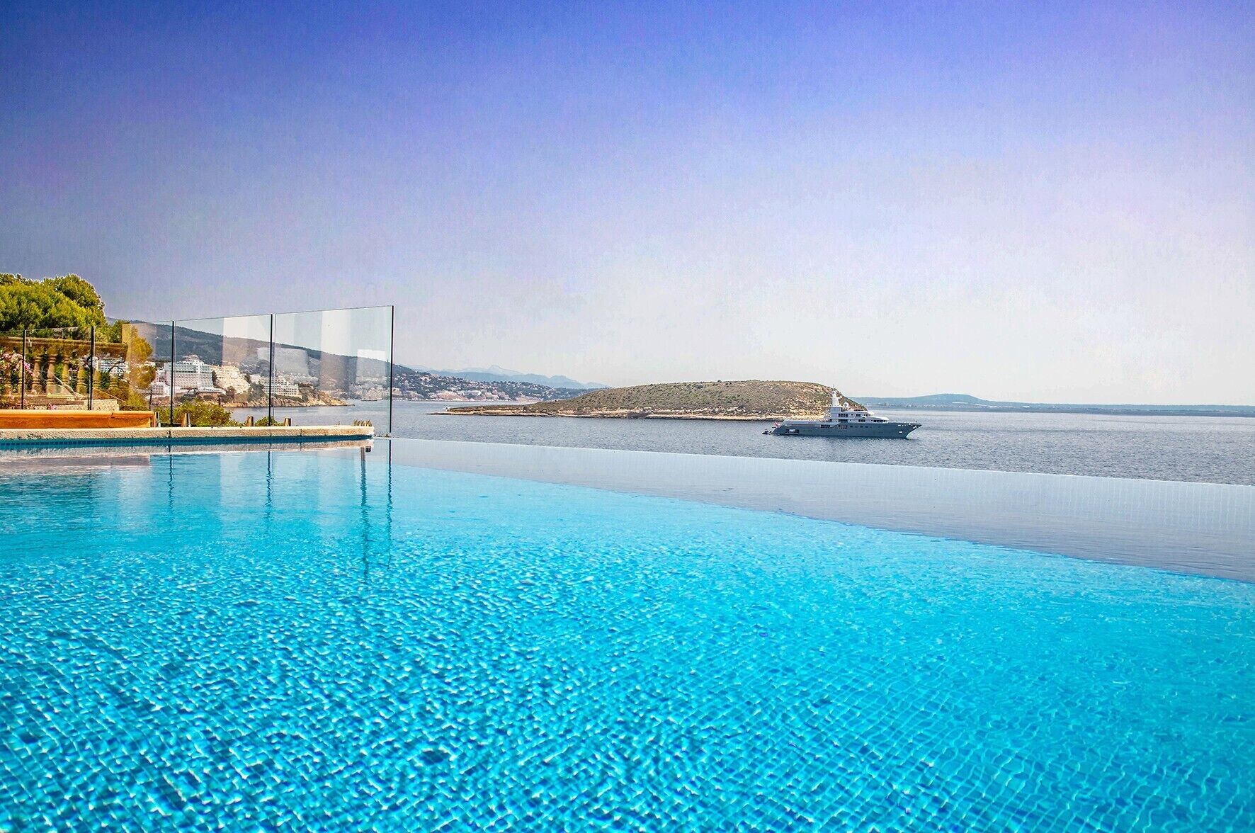 Uma piscina infinita azul cristalina com vista para o mar e um iate ao longe.