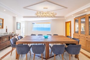 Une salle à manger avec une grande table en bois, six chaises grises, un bol bleu, un lustre et une vue sur la mer par une grande fenêtre.