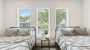 Ein Schlafzimmer mit zwei Betten, einem kleinen Tisch mit Lampe und drei Fenstern mit Jalousien.