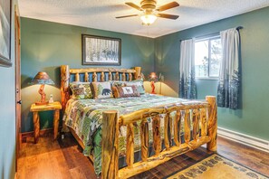 Une chambre à coucher avec une tête de lit en bois, un lit recouvert d’une literie à motifs, une table de chevet avec une lampe, un ventilateur de plafond et une fenêtre avec des rideaux.