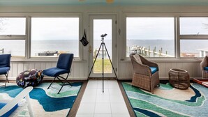 A sunroom with a view of the sea, featuring a ceiling fan, a tripod, and seating areas.
