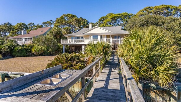 A coastal house with a wooden deck leading to a covered porch, surrounded by lush vegetation.