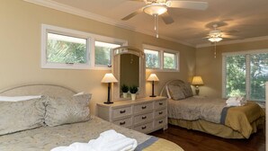 A bedroom with two beds, a dresser, two lamps, a ceiling fan, and large windows.