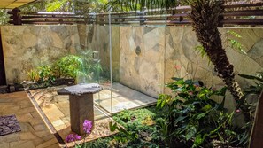 Un espace de douche clos par des parois de verre, doté d’un socle en pierre, entouré de plantes tropicales et d’un mur en pierre.