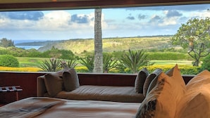 Ein Zimmer mit großem Fenster, das einen angelegten Garten und den Blick aufs Meer bietet.
