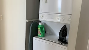 Une petite buanderie avec un lave-linge et un sèche-linge blancs, une bouteille verte de lessive et un fer à repasser noir posé sur le sèche-linge.