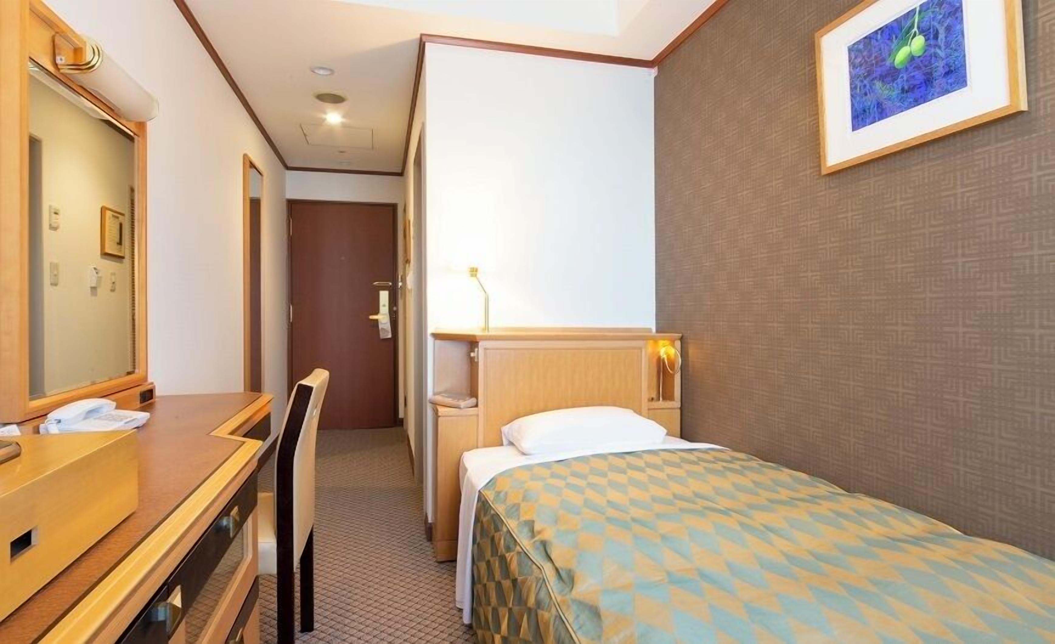 Een hotelkamer met een bed, een bureau, een stoel, een spiegel, een nachtkastje, een lamp en een lijst met een foto aan de muur.