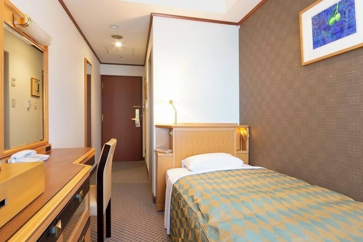A hotel room with a bed, a desk, a chair, a mirror, a bedside table, a lamp, and a framed picture on the wall.