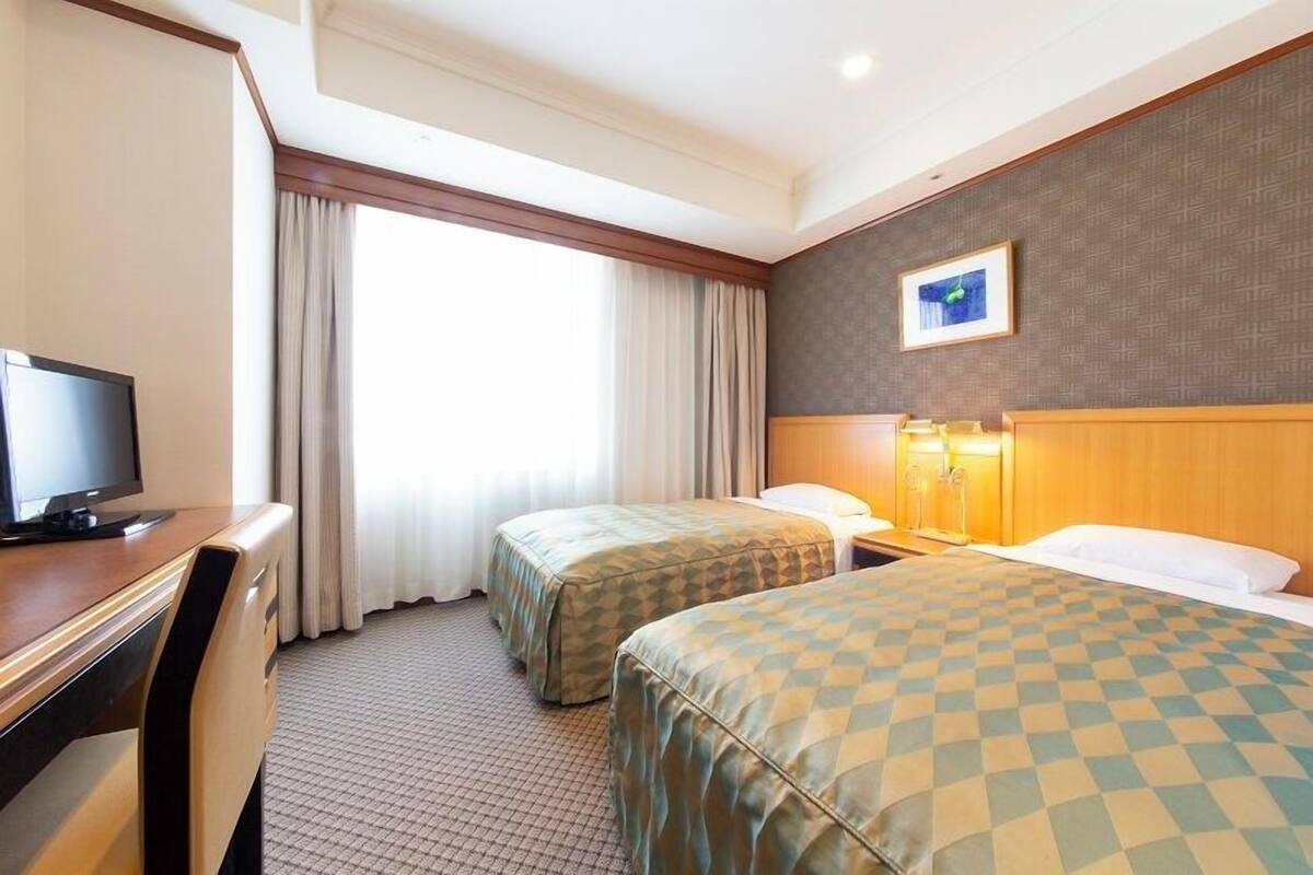 A hotel room with two beds, a desk with a television, a window with curtains, and a framed picture on the wall.