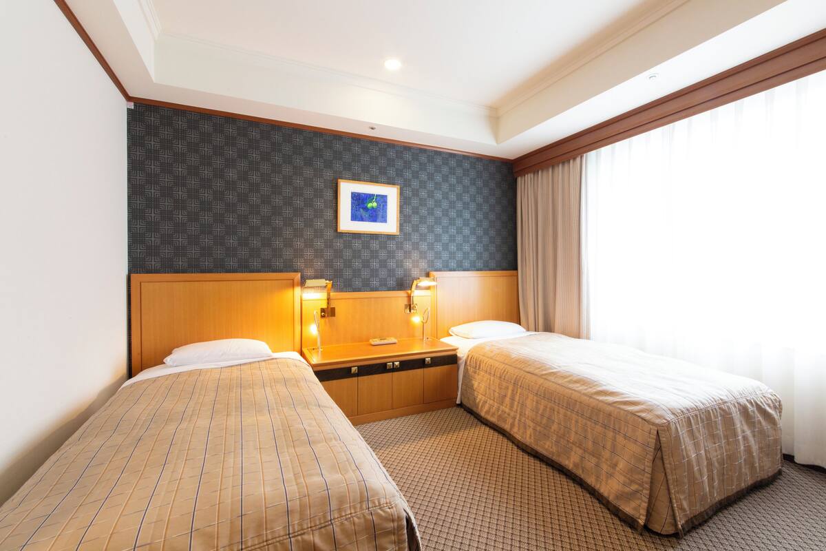 A hotel room with two beds, a headboard, a nightstand, a framed picture, and a window with curtains.