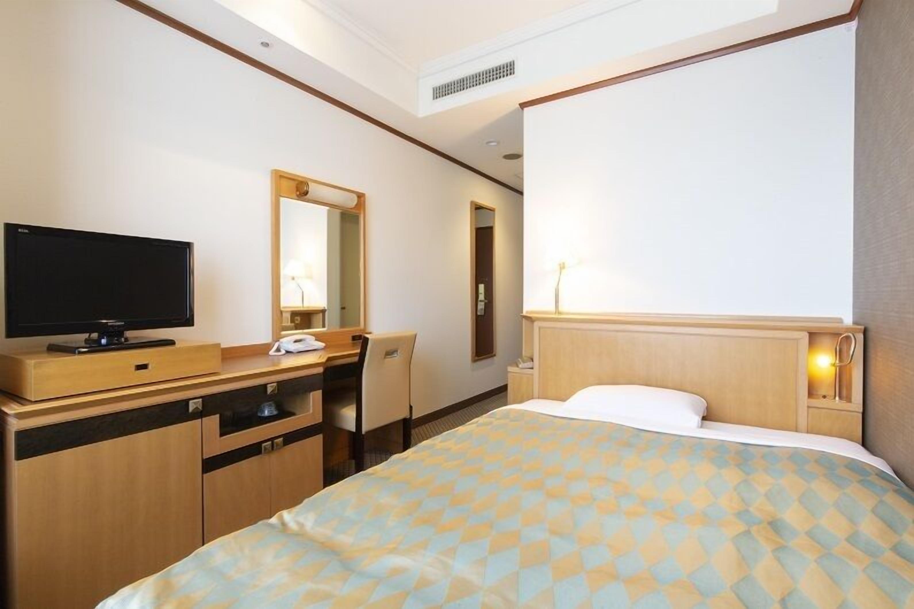 A hotel room with a bed, a desk with a television, a mirror, and a bedside table with a lamp.