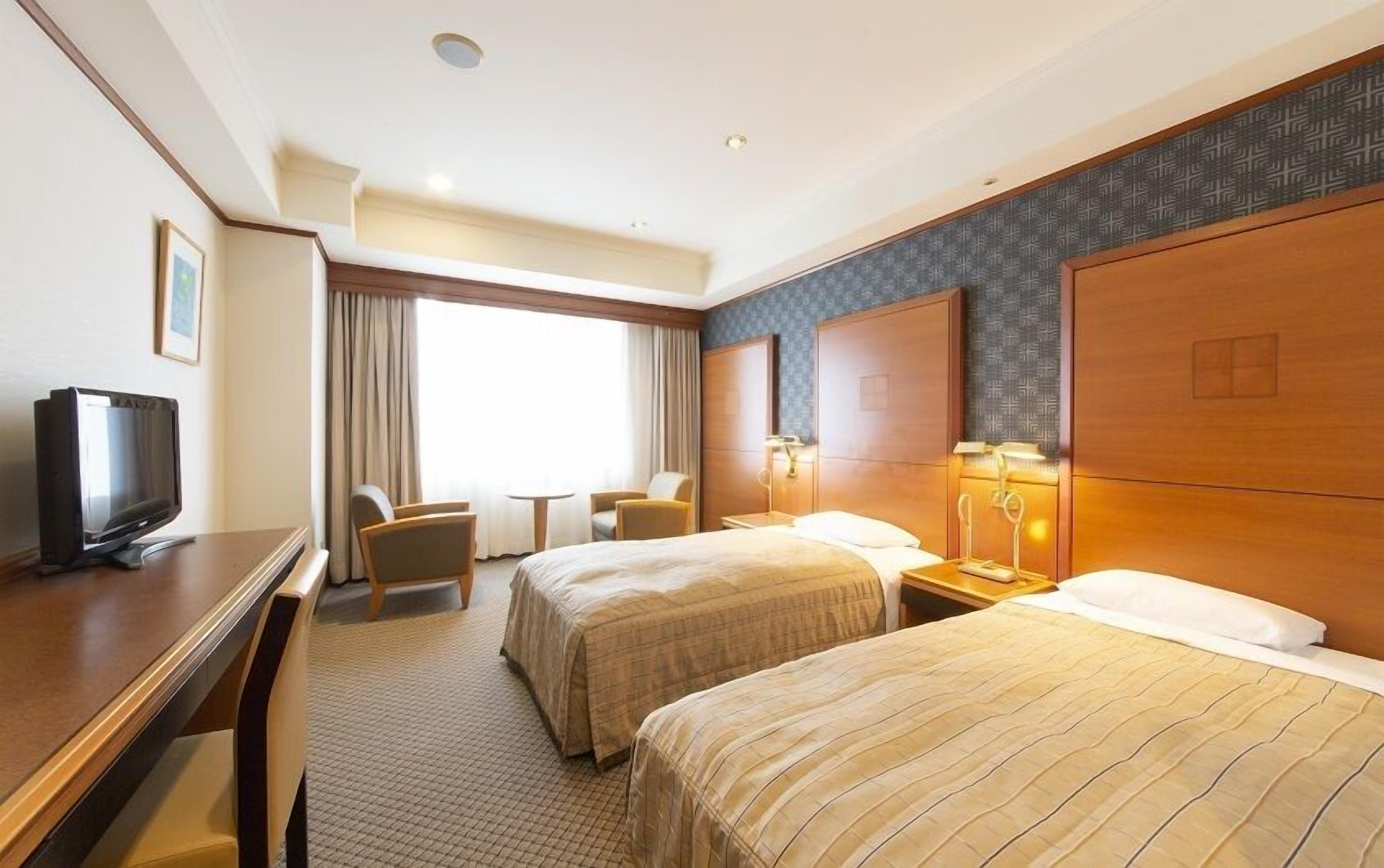 A hotel room with two beds, a desk with a television, a small table, and a window with curtains.