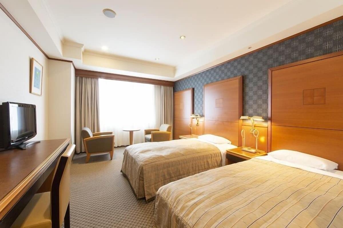 Habitación de hotel con dos camas, un escritorio con televisor, una mesita y una ventana con cortinas.