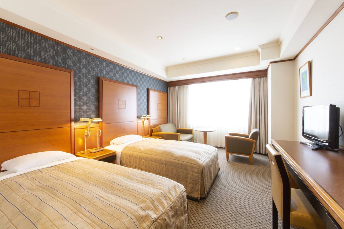 A hotel room with two beds, a desk, a television, and a window with curtains.