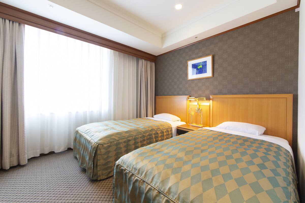 A hotel room with two beds, a large window with curtains, a framed picture on the wall, and a nightstand with a lamp.