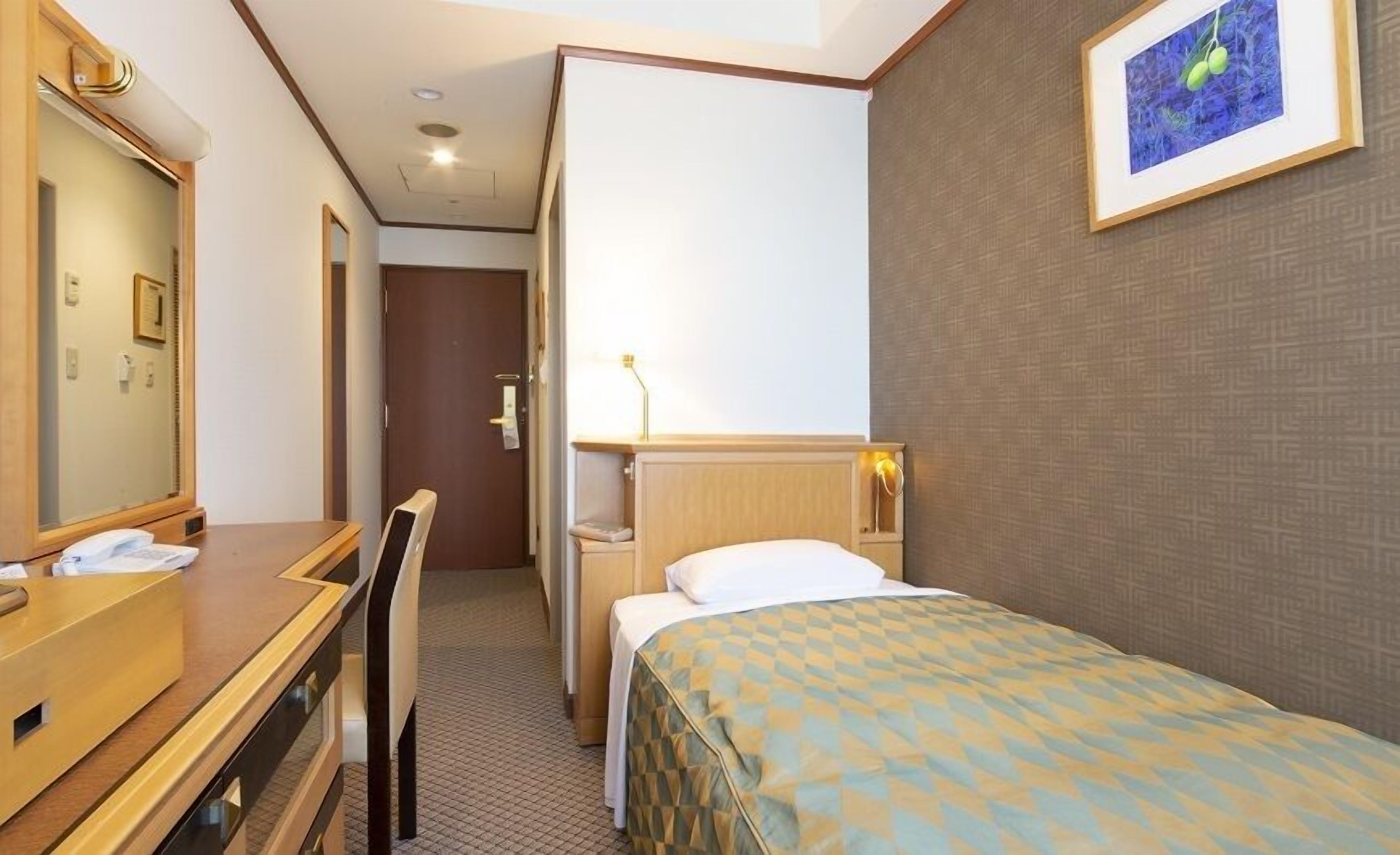 A hotel room with a bed, a desk, a chair, a mirror, a bedside table, a lamp, and a framed picture on the wall.