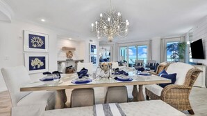 A spacious dining area with a glass table, white upholstered chairs, and a chandelier.