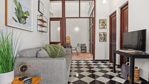 Ein Wohnzimmer mit grauem Sofa, einem Couchtisch und einem TV-Schrank. Der Boden ist kariert und eine Tür führt in einen anderen Raum.
