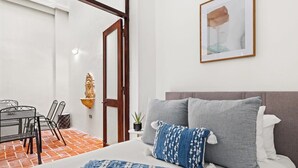 Ein Schlafzimmer mit Bett, Kissen, einem Nachttisch, einem gerahmten Bild, einem Essbereich mit Stühlen und Tisch sowie einer Topfpflanze.