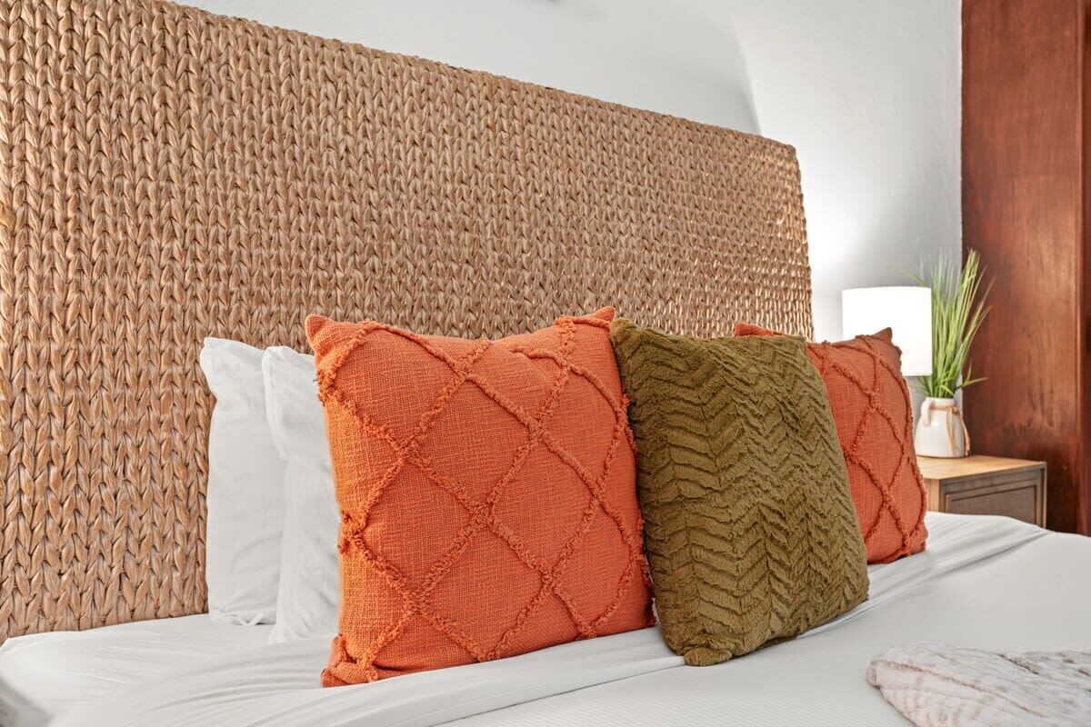 Cama com cabeceira de fibra trançada, almofadas brancas e laranja, e um travesseiro verde com textura.