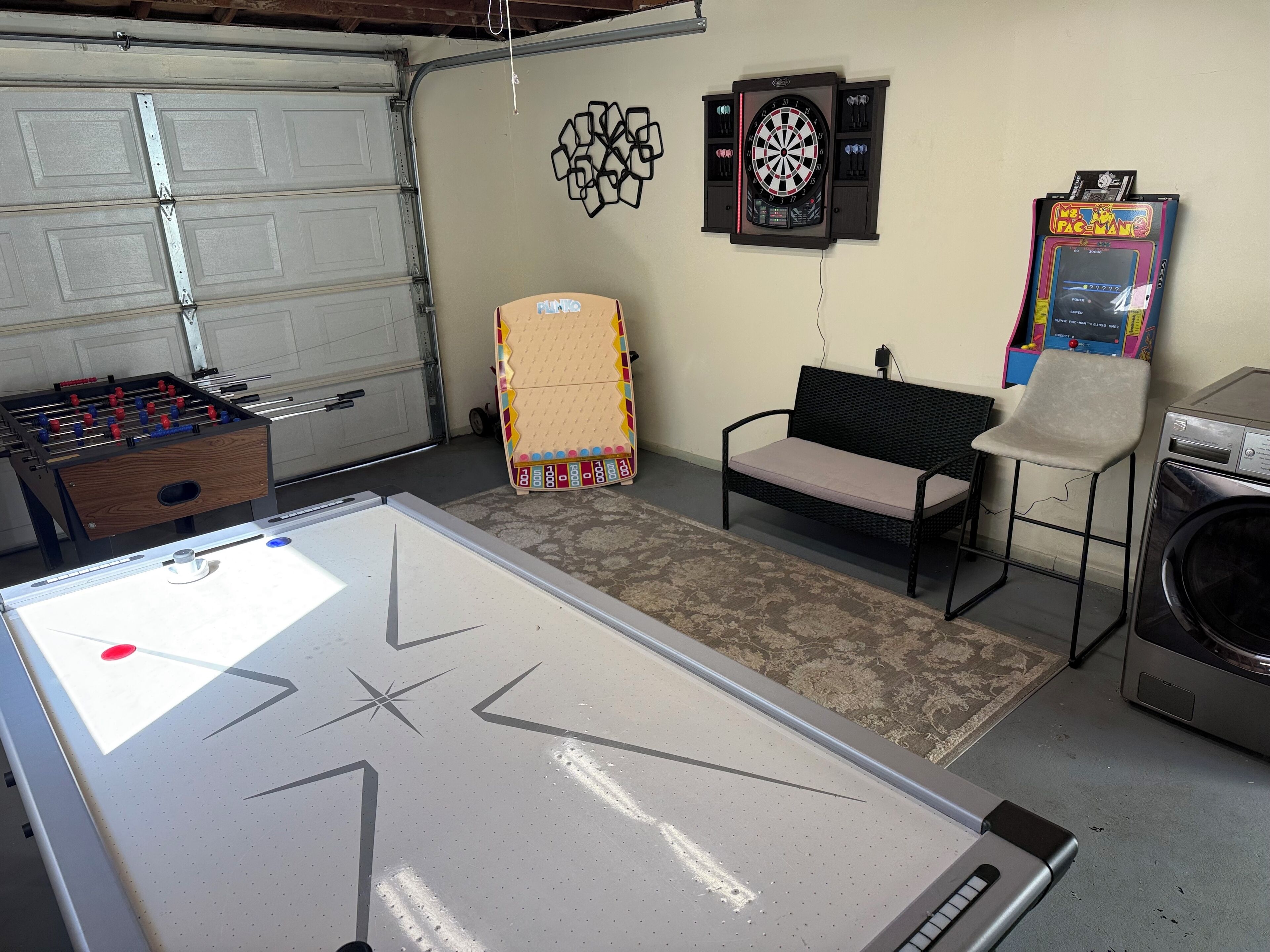 A recreational room with an air hockey table, a foosball table, a dartboard, a bench, and a washing machine.