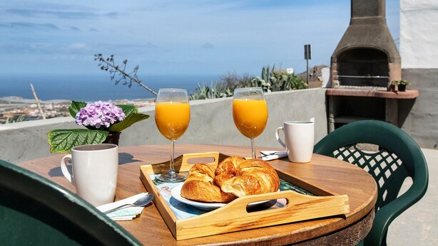 Bandeja de desayuno con cruasanes, dos vasos de zumo de naranja y una taza blanca sobre una mesa de madera con vistas al mar y un horno de ladrillo al fondo.
