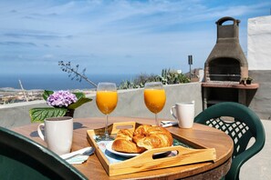 Ein Frühstücksbrett mit Croissants, zwei Gläser Orangensaft und einer weißen Tasse auf einem Holztisch. Im Hintergrund Meerblick und ein Backofen.