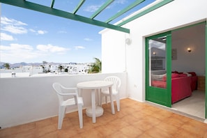 Un balcone con tavolo e sedie bianche, una porta con cornice verde e vista su edifici e montagne.