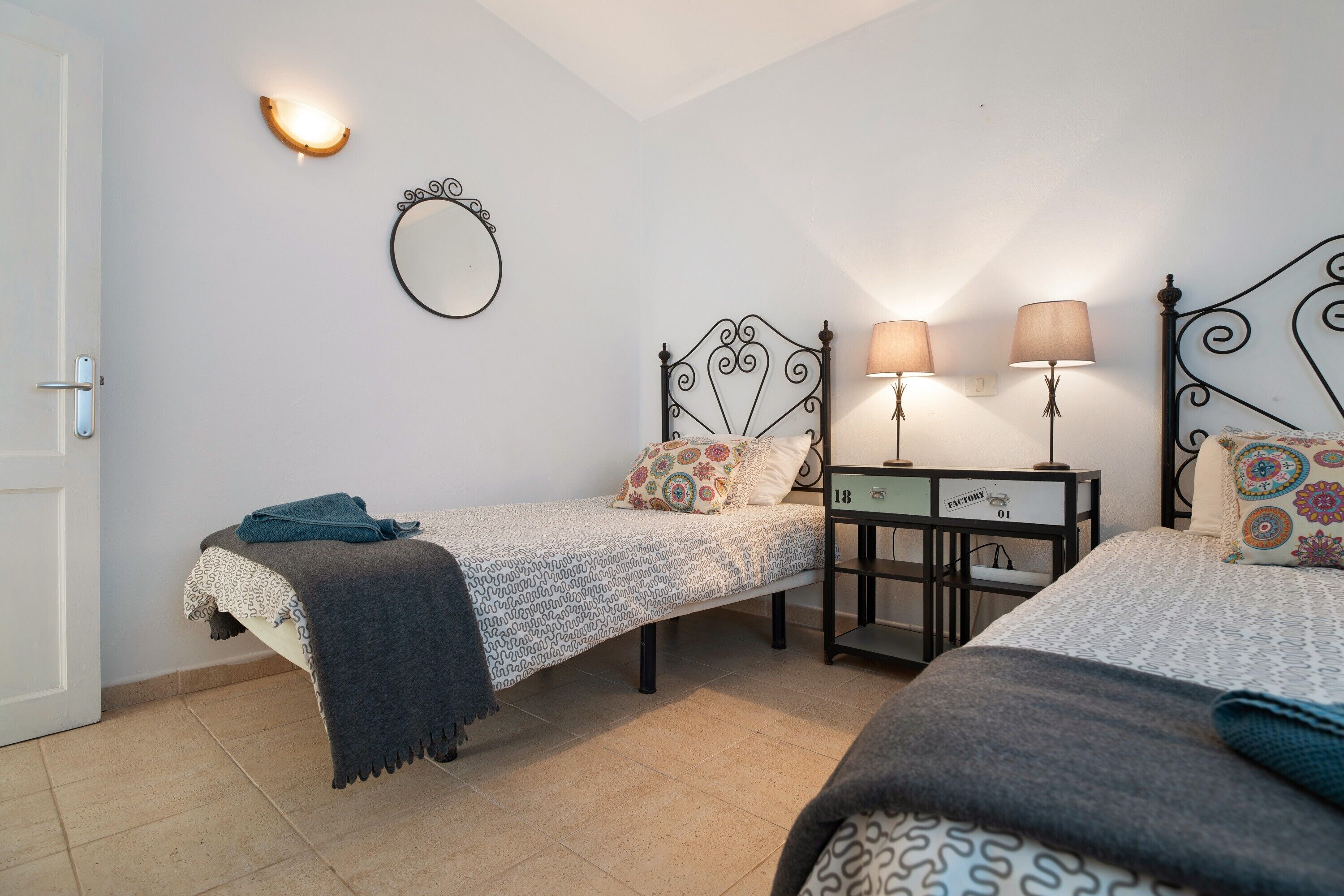 A bedroom with two beds, a nightstand, a mirror, and wall-mounted lamps.