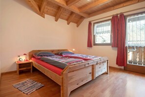 A wooden bed with a headboard, a nightstand with a lamp, a window with curtains, and a wooden floor.