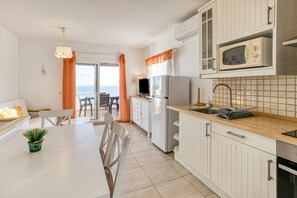 Una cucina moderna con mobili bianchi, un forno a microonde e un frigorifero. C'è un'area pranzo con tavolo e sedie, e vista sull'oceano attraverso una porta di vetro.