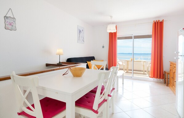 Un coin repas avec une table et des chaises blanches, un saladier posé sur la table, et une vue sur la mer par une porte vitrée.