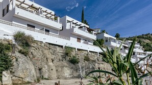 Un edificio costero con varios balcones, paredes exteriores blancas y un paisaje de colinas rocosas.