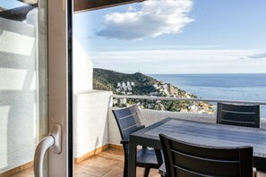 Un balcon avec une table et des chaises, offrant une vue sur la côte, avec des bâtiments et des collines en arrière-plan.